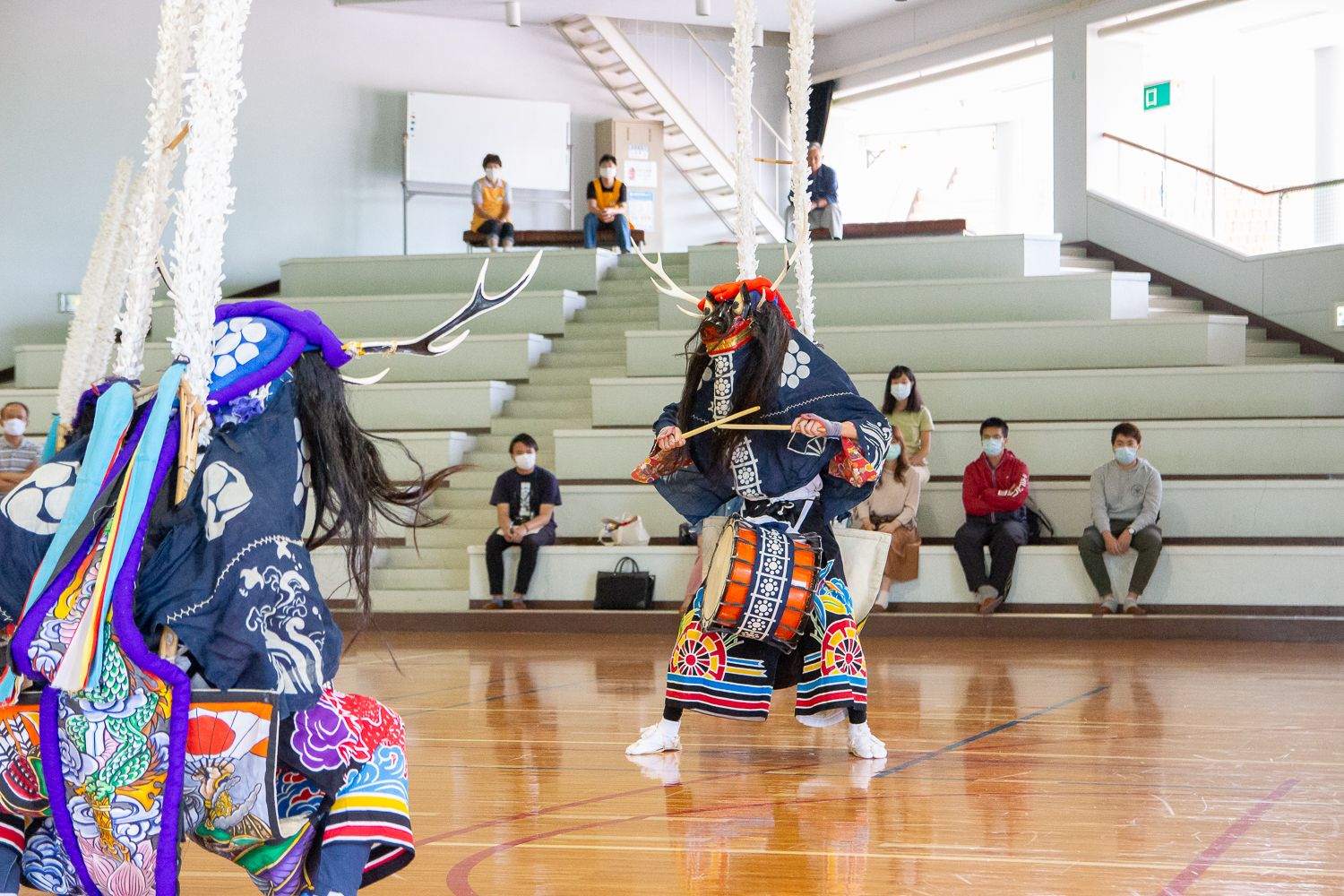 体育館のような室内で、カラフルな衣装と鬼のような面をつけた二人の舞い手が太鼓を叩きながら伝統的な踊りを披露しており、階段状の観客席にはマスクを着けた参加者たちが静かに見守っている写真