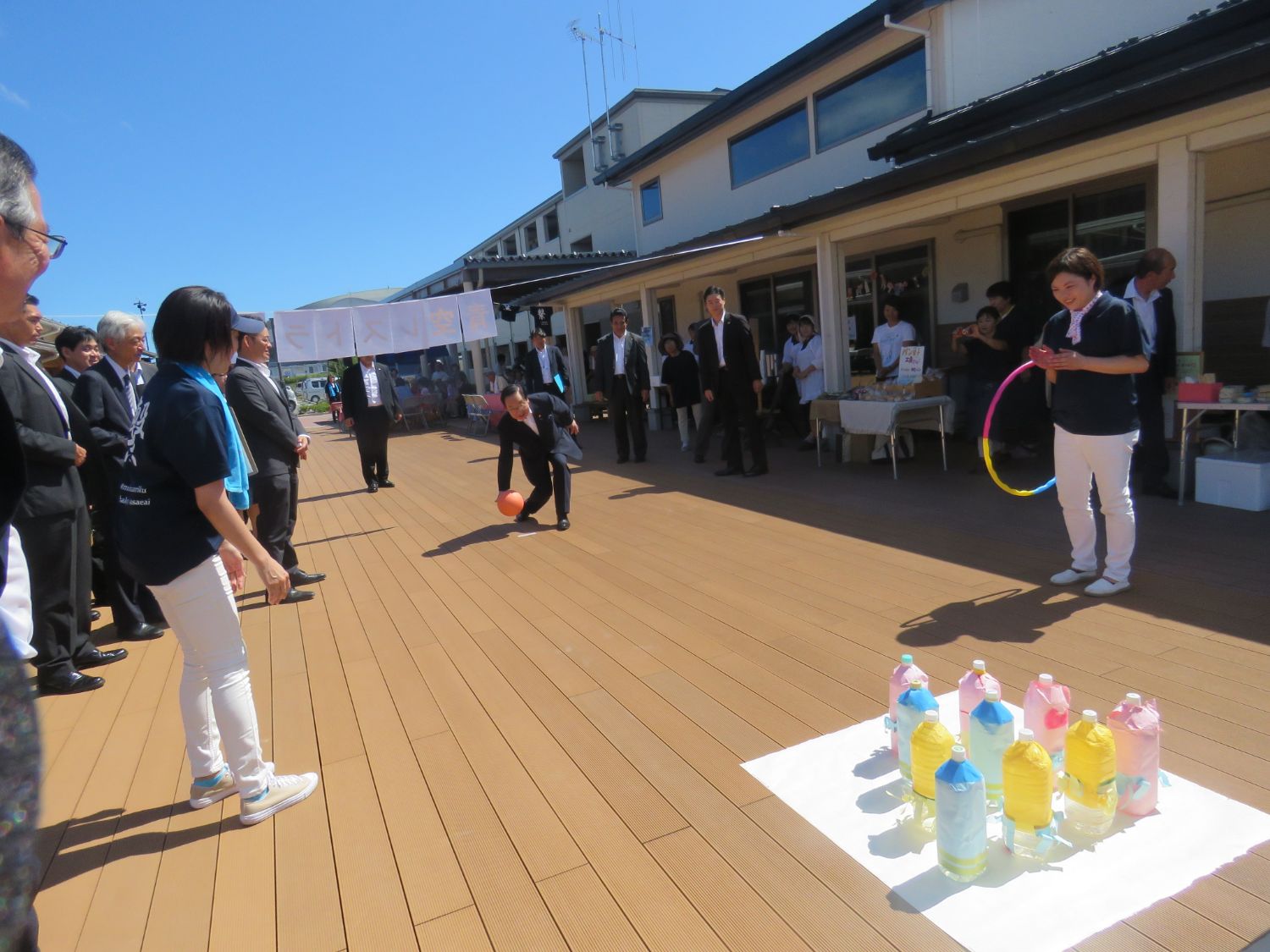 晴天の屋外ウッドデッキで、スーツ姿の男性がオレンジ色のボールを投げてペットボトルを使ったボウリングのようなゲームに挑戦しており、周囲では見守る人々が笑顔で立ち並び、和やかな雰囲気の中でイベントが進行している写真
