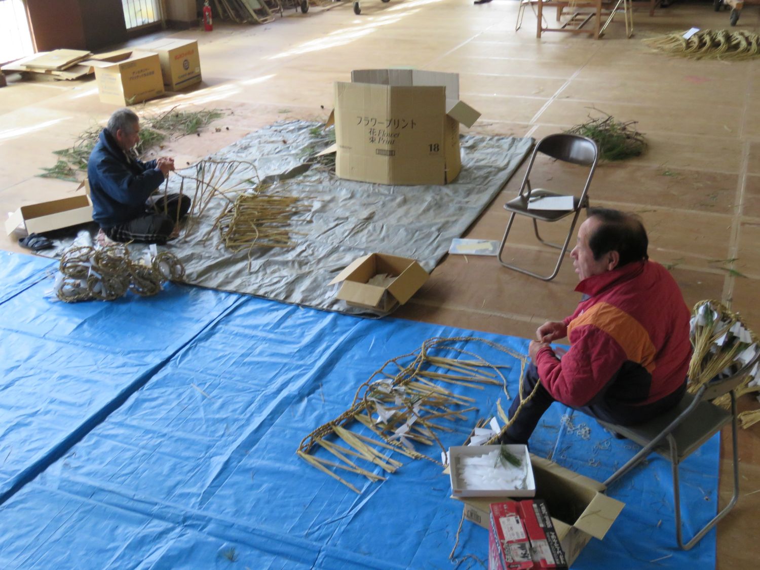 ブルーシートの上にパイプ椅子に座りしめ縄を編んでいる山内さん、白いシートの上であぐらを組んでしめ縄を編んでいる佐藤さんの写真