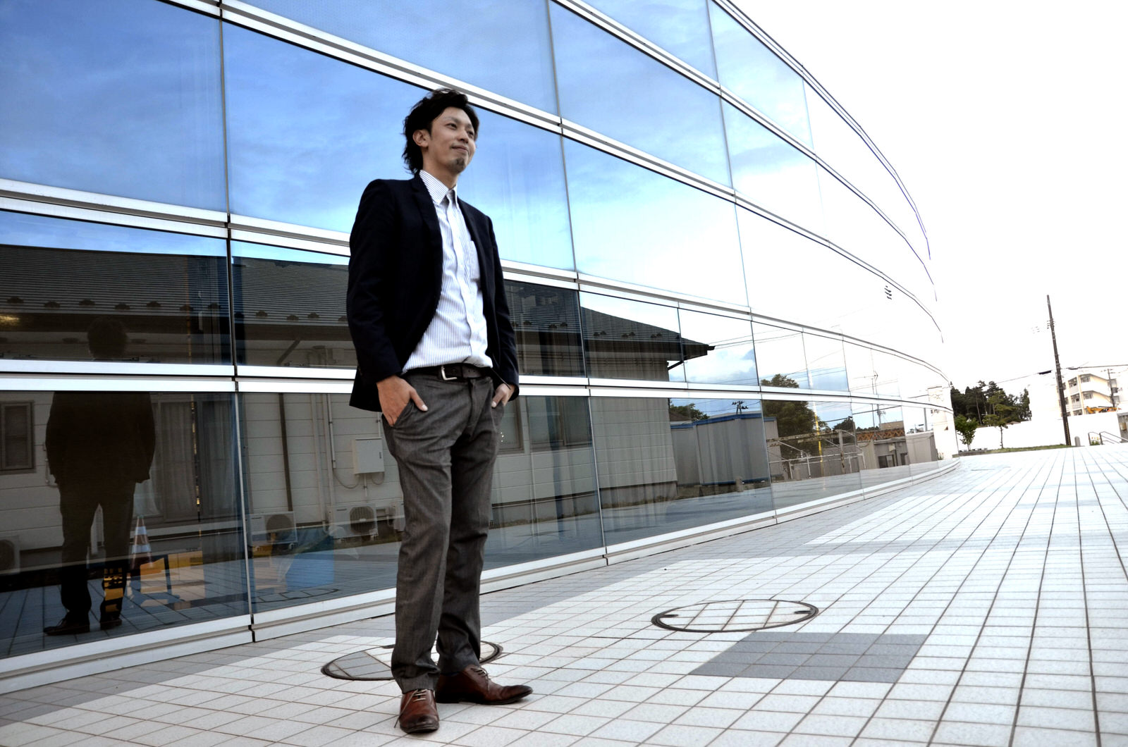 近代的なガラス張りの建物の前にスーツ姿の井尻さんが立ち、手をポケットに入れて穏やかに微笑んでいる写真