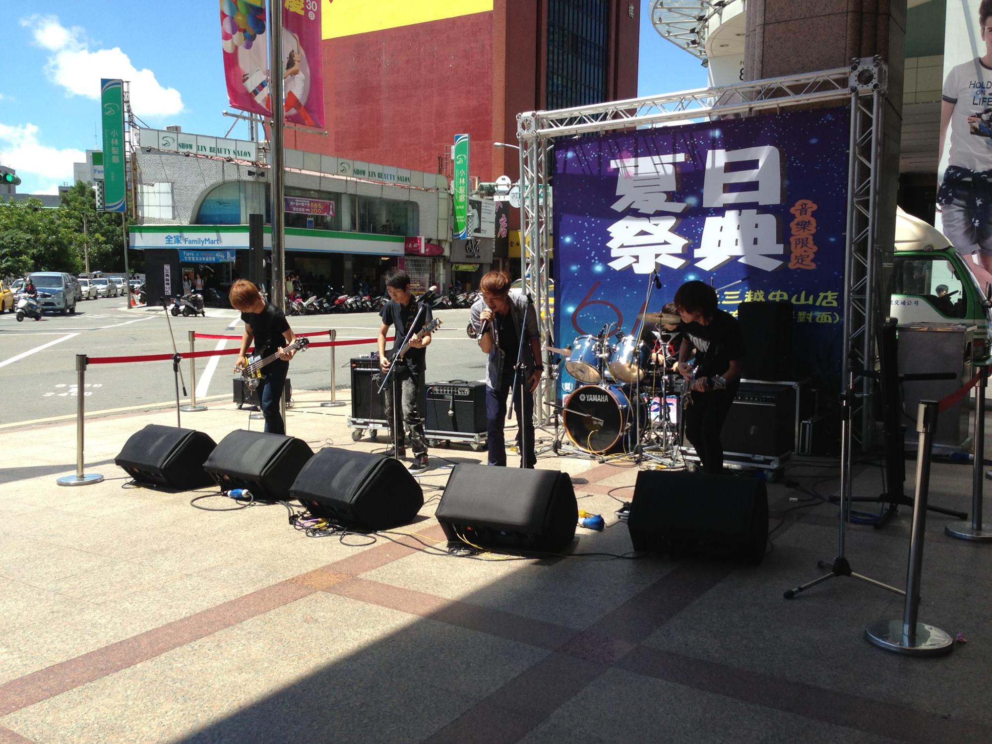 「夏日祭典」と書かれた大きなイベント用のバックボードが設置された街中の広場で、若者のバンドがドラムやギターなどでライブ演奏をしている写真
