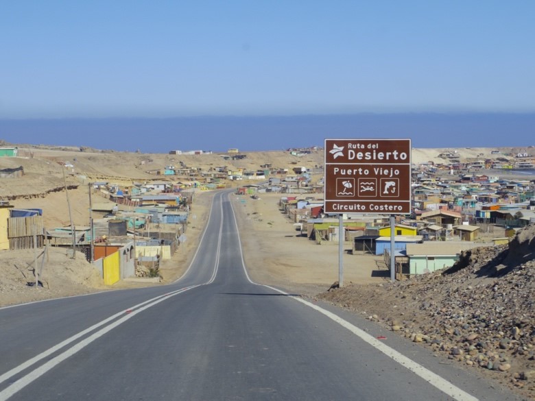 舗装された道が小さな砂漠の町へと続き、右側には「Ruta del Desierto（砂漠の道）」と書かれた観光案内標識が立っている写真