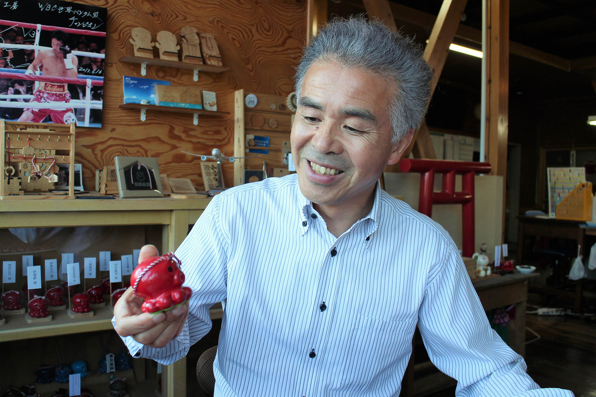 工房のような室内で阿部忠義さんが笑顔で手のひらサイズの赤いタコの置物を見つめている写真