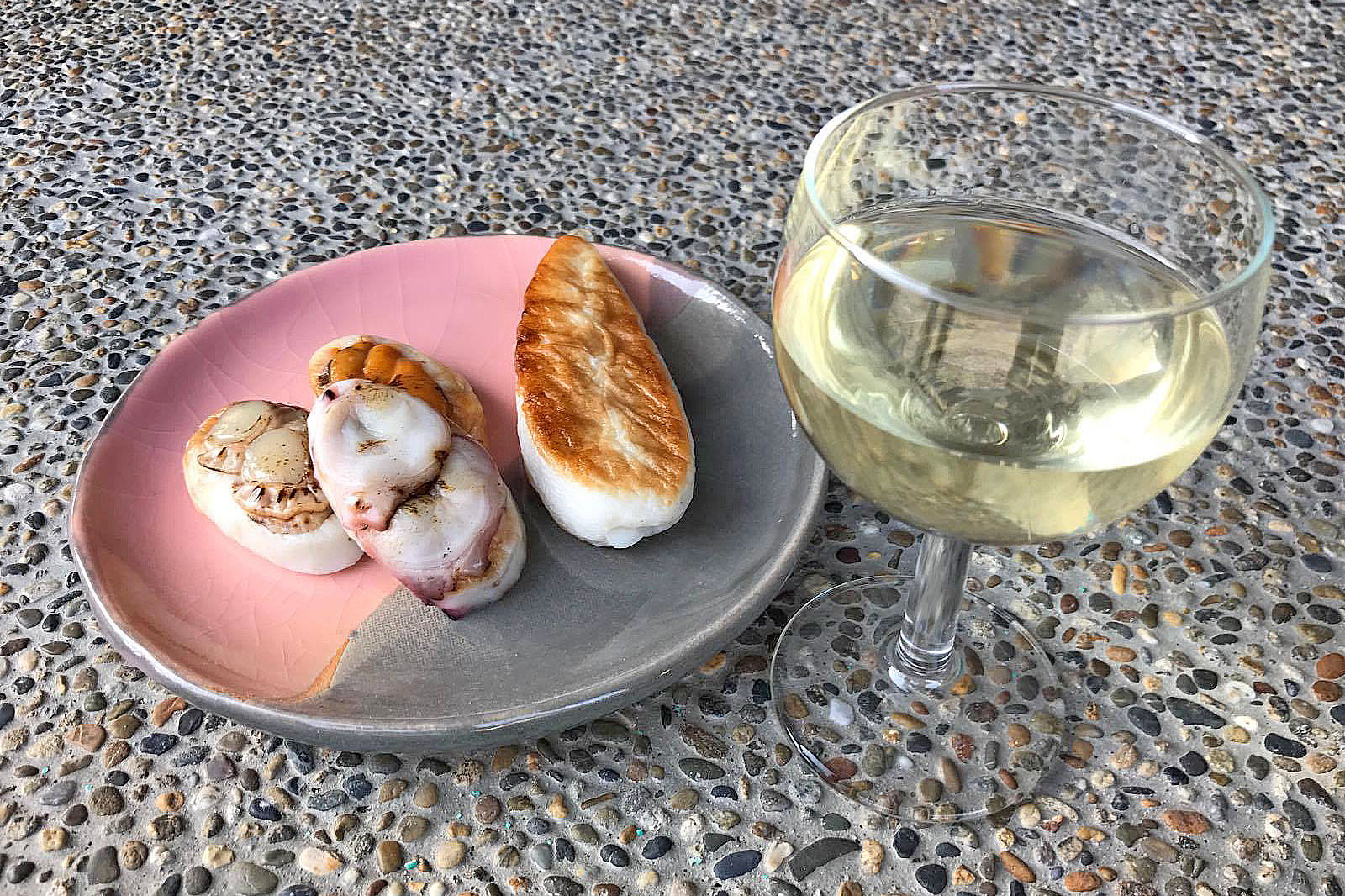 ピンクとグレーの陶器皿に盛られた焼き蒲鉾やホタテなどのおつまみと、白ワインが入ったグラスが並ぶ写真