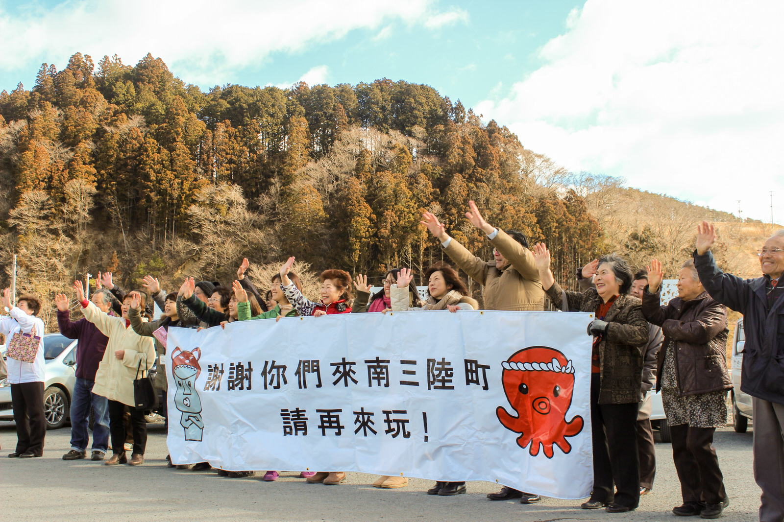 森を背景にした港で地元の人々が横断幕を持って手を振りながら、民泊体験学習に参加した外国人に感謝と再訪を呼びかけている写真