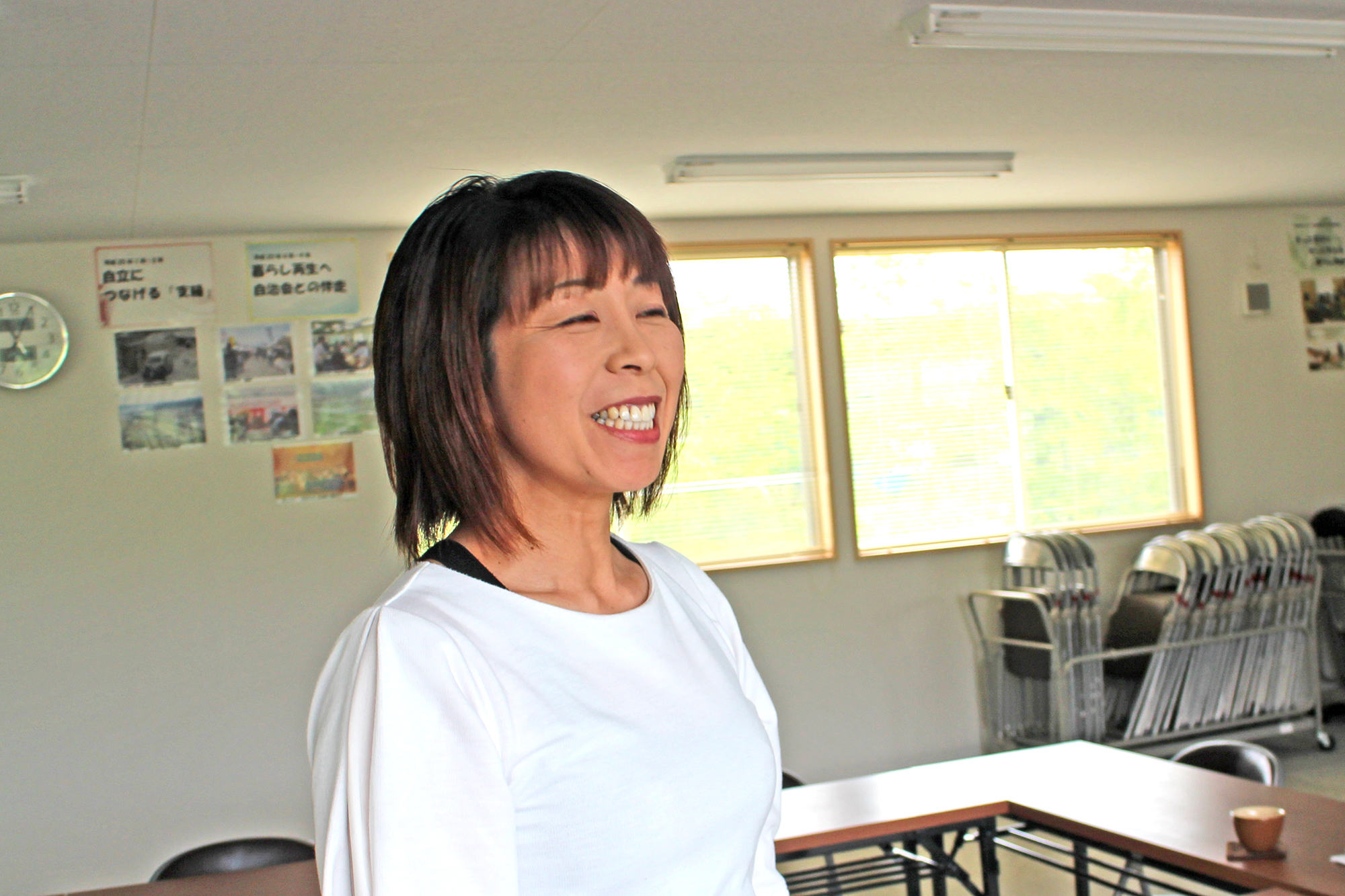 長机と椅子が並んだ室内で高橋さんが少し右を向いて笑顔を見せている写真