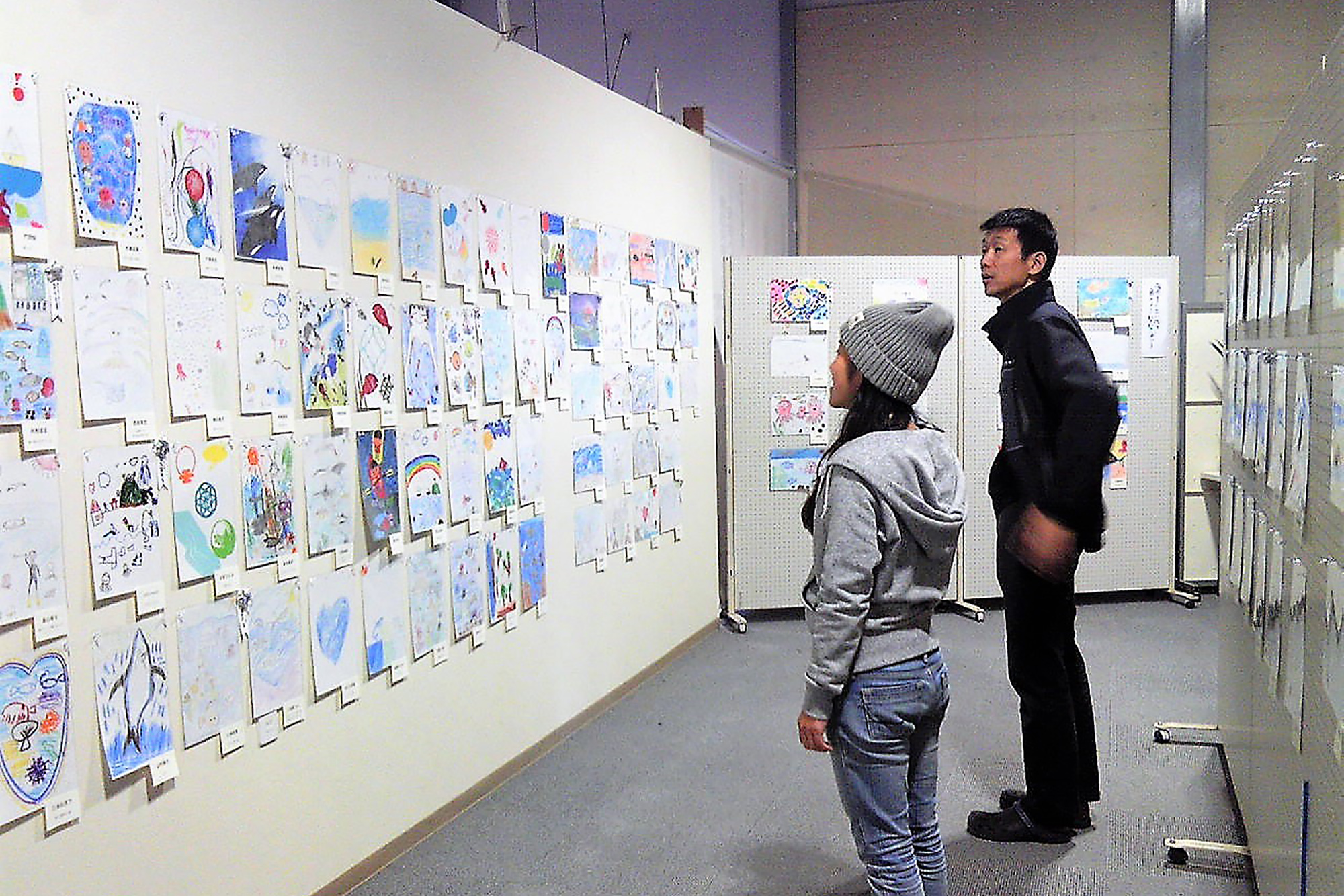 室内の展示スペースで、2人の来場者が壁一面に並べて展示された子どもたちの色とりどりの絵を静かに鑑賞している写真