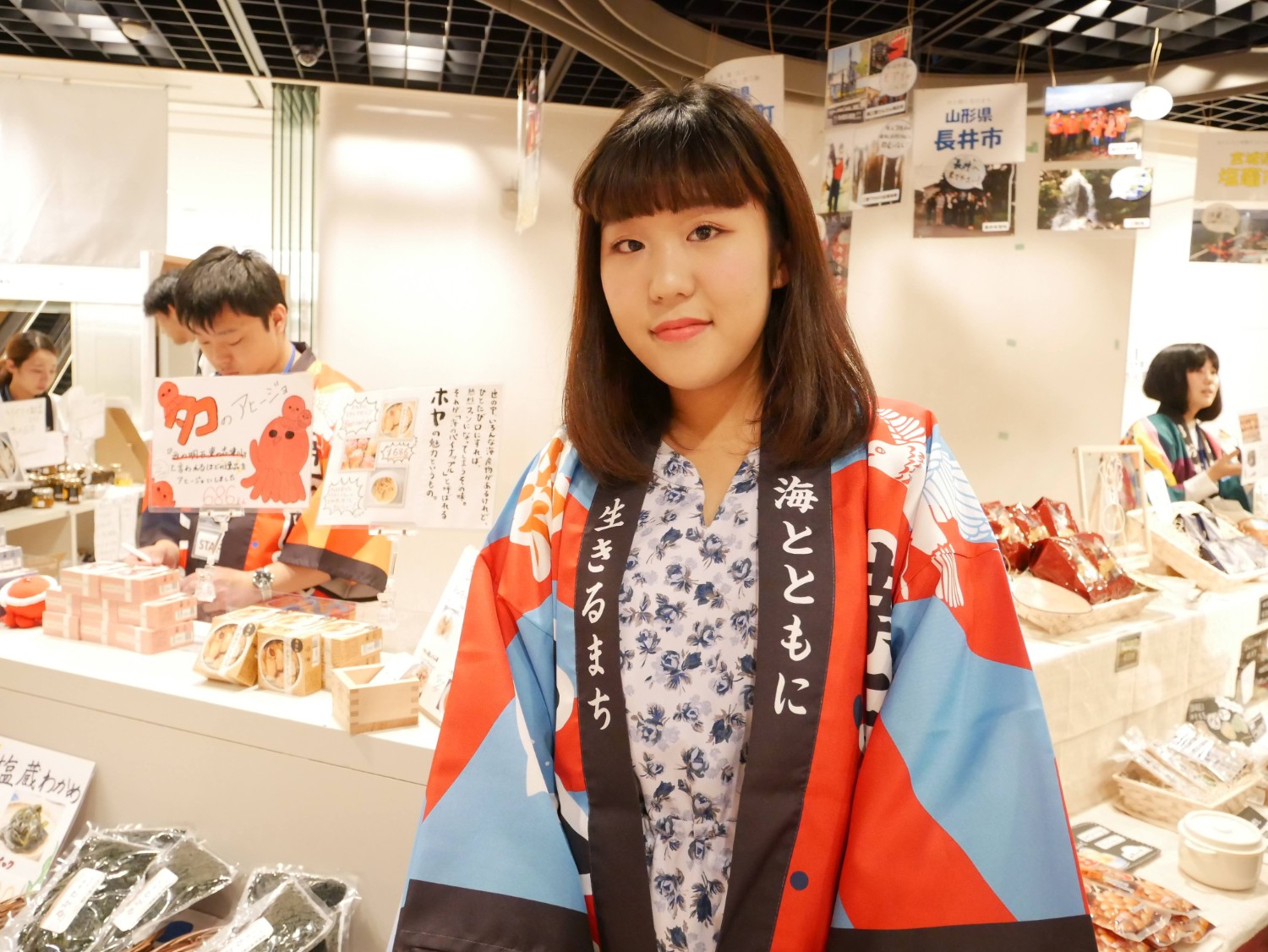 「海とともに生きるまち」と書かれた文字が目立つカラフルな法被を着た若い女性が、物産展のブース前で穏やかな表情を見せており、背後には「タコのアヒージョ」などの手描きポップや海産物のパッケージ商品が並ぶにぎやかな売り場が広がっている写真