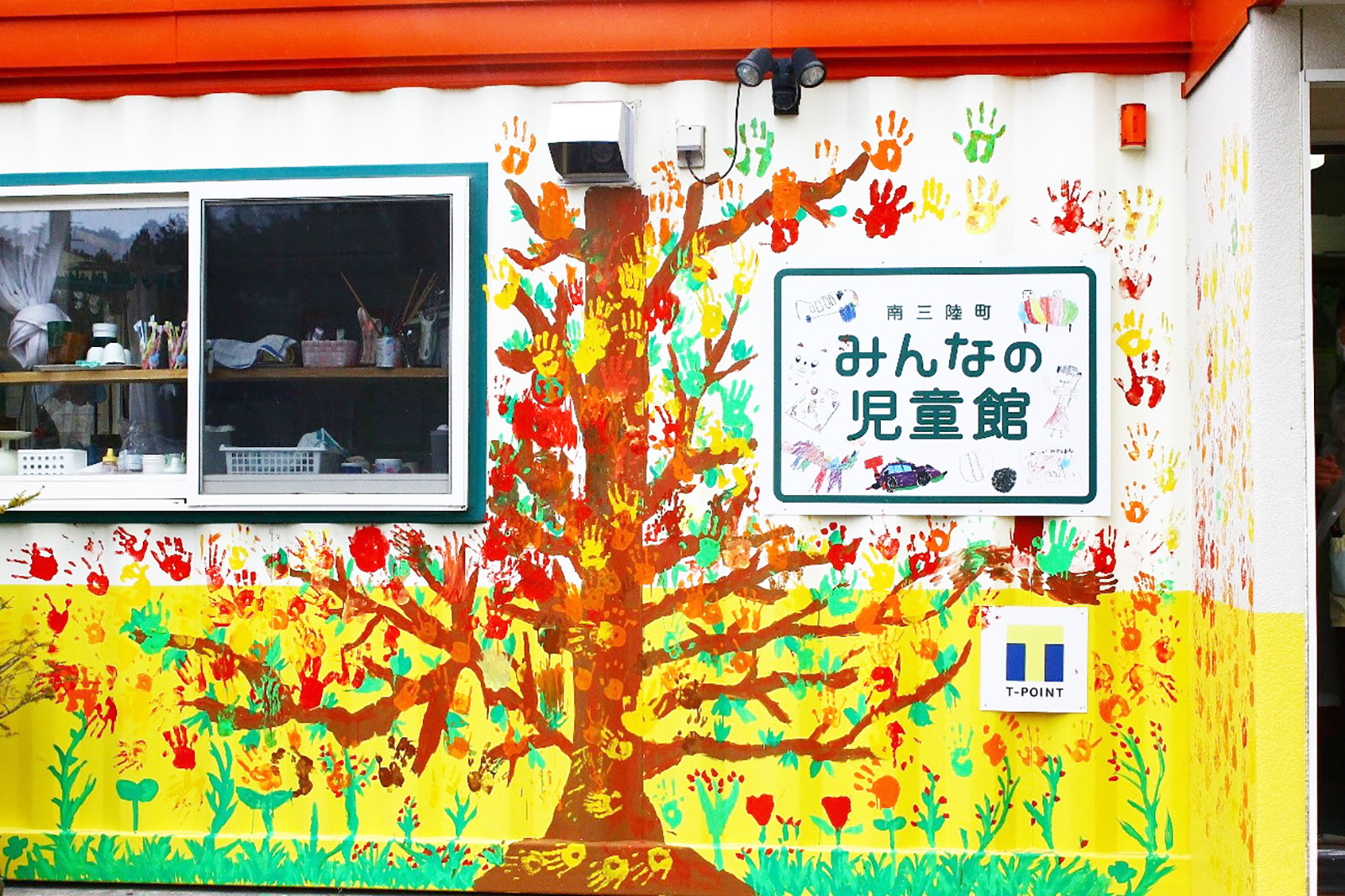 「南三陸町みんなの児童館」と書かれた看板が設置されたコンテナハウスの外壁一面に色鮮やかな手形アートの大きな木が描かれている写真