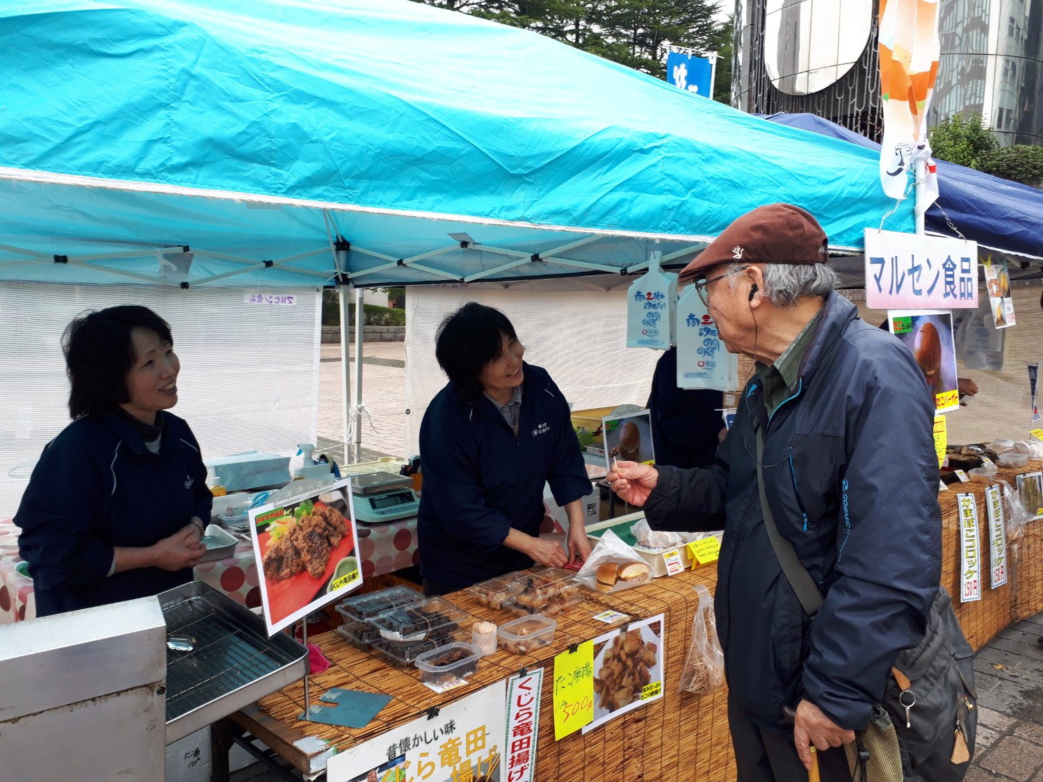 商品の写真が掲示された机にコロッケや揚げ物などの食品が並んでいる「マルセン食品」の看板が掲げられたブースで、試食をしている年配の男性客に、女性店員が笑顔で接客している様子の写真