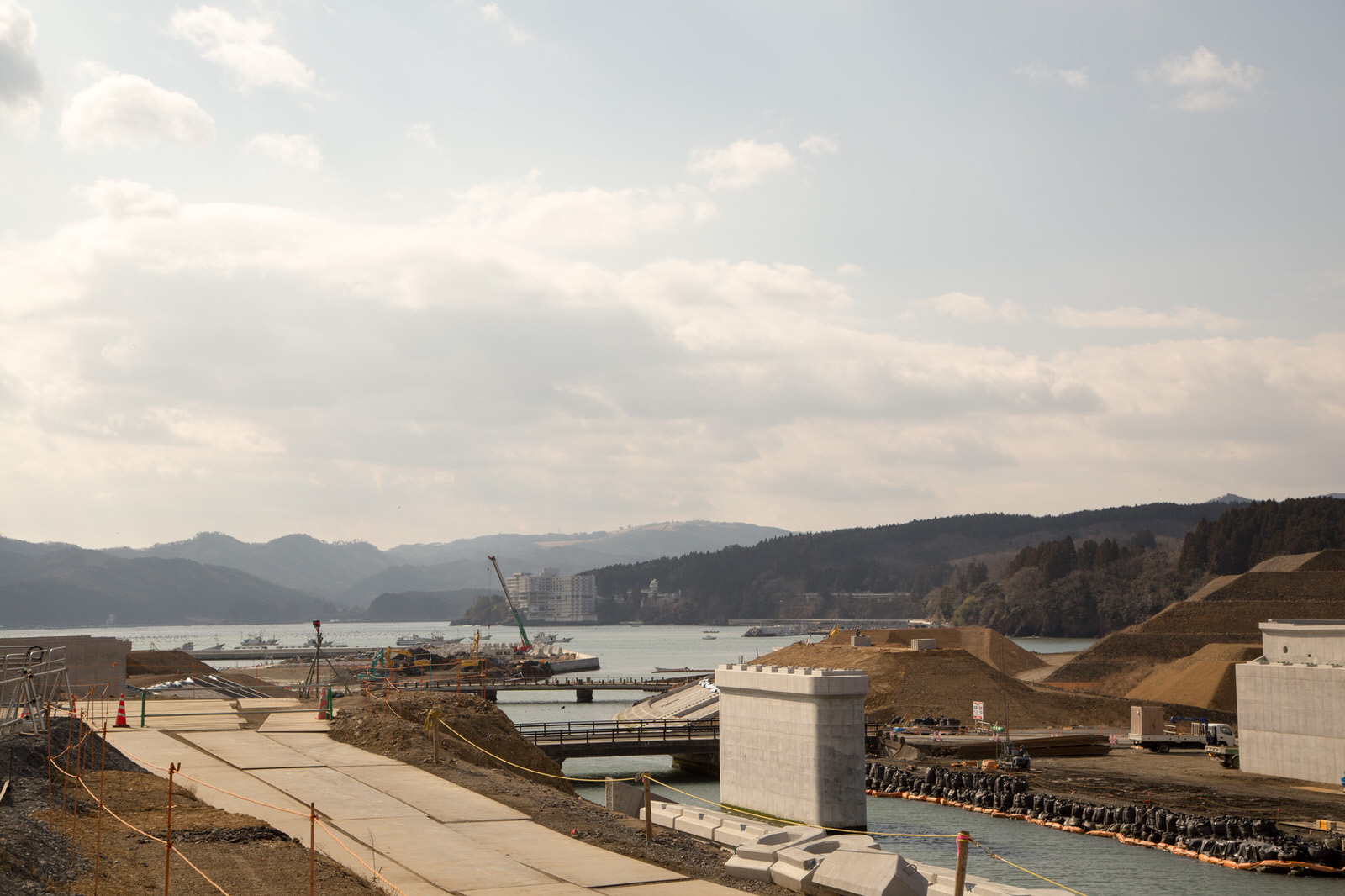 山々に囲まれた海沿いの場所で、複数の重機やクレーンが稼働していて、護岸工事が行われている様子が見え、奥には港町のような景色が広がっている、震災復興のための工事現場の写真