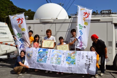 のぼり旗を持ち、受賞した作品が掲示された大きな模造紙を広げ持ったスタッフたちの後ろで、2人の受賞者が賞状を持って笑顔で並んでいる記念写真