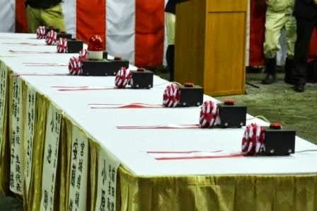 白い布が敷かれた長いテーブルに代表者の所属先と名前が記載された垂れ幕が掲げられ、それぞれに赤白のリボンが飾られている赤いボタンが付いた黒い箱がいくつも並べられた写真