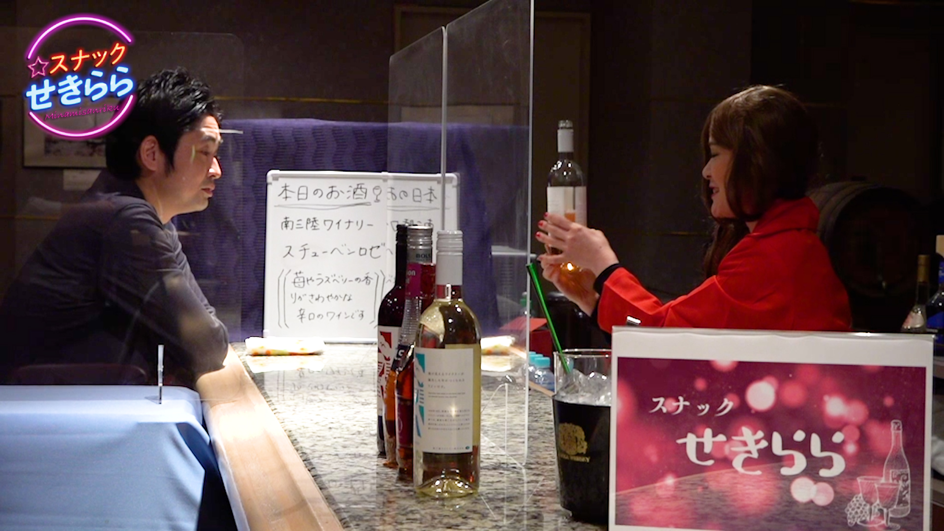 カウンター越しに男性と女性が向かい合って座り、男性は真剣な表情で女性を見つめ、女性は白ワインの瓶を手に持ち、説明している様子の写真