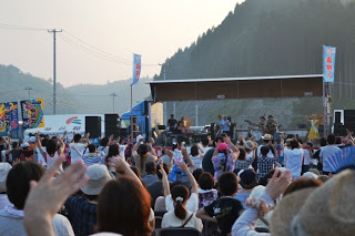 夕暮れ時の野外ステージでバンドが演奏し、それに合わせて大勢の観客が手を振りながら盛り上がっている様子を写した写真