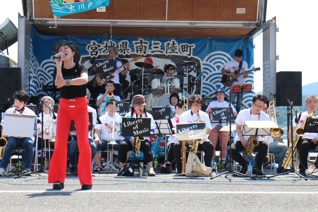 「宮城県南三陸町」と書かれた青い横断幕が掲げられている屋外の特設ステージで、後方ではサックスやトランペット、ギター、ドラムなど複数の楽器を持つバンドの演奏に合わせて、黒い帽子と黒いシャツ、赤いズボンを着た女性ボーカルが情熱的に歌っている写真