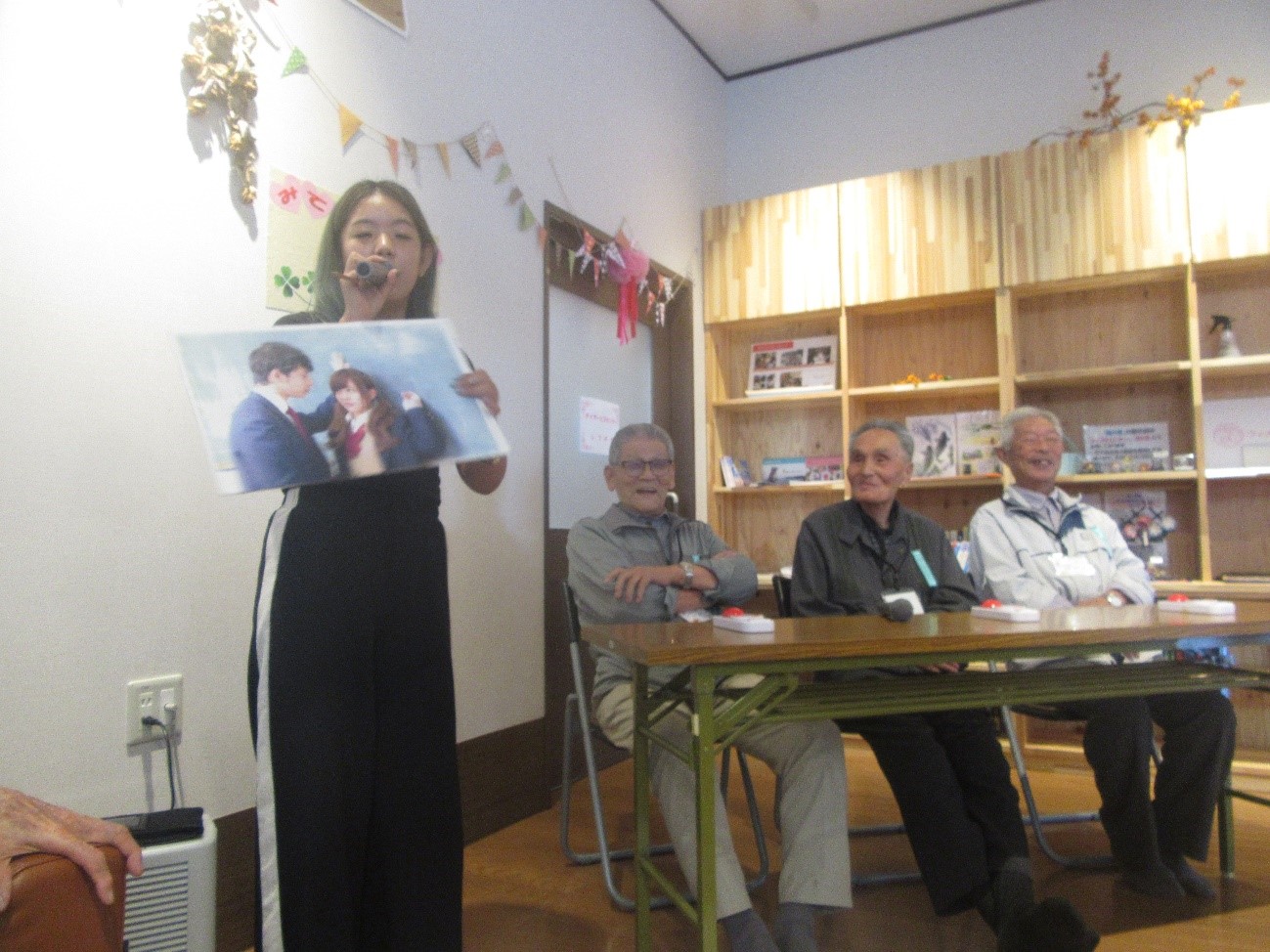 女性司会者が制服を着た男子学生が女子学生に壁ドンを行っている場面写真のパネルを持って説明しており、それを見て高齢の男性三人が笑顔で反応しているクイズ中の写真