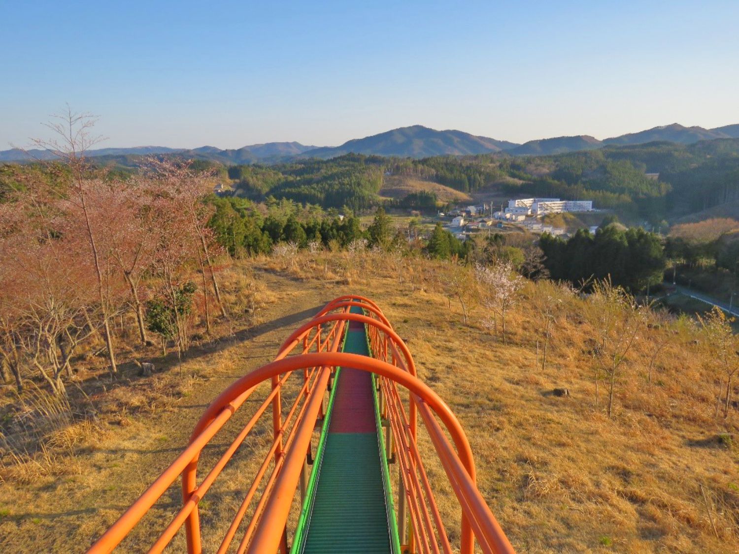 丘の上からオレンジ色の手すりの長い滑り台が伸び、見下ろす先には山々と町並みが広がる夕暮れ時の展望の写真
