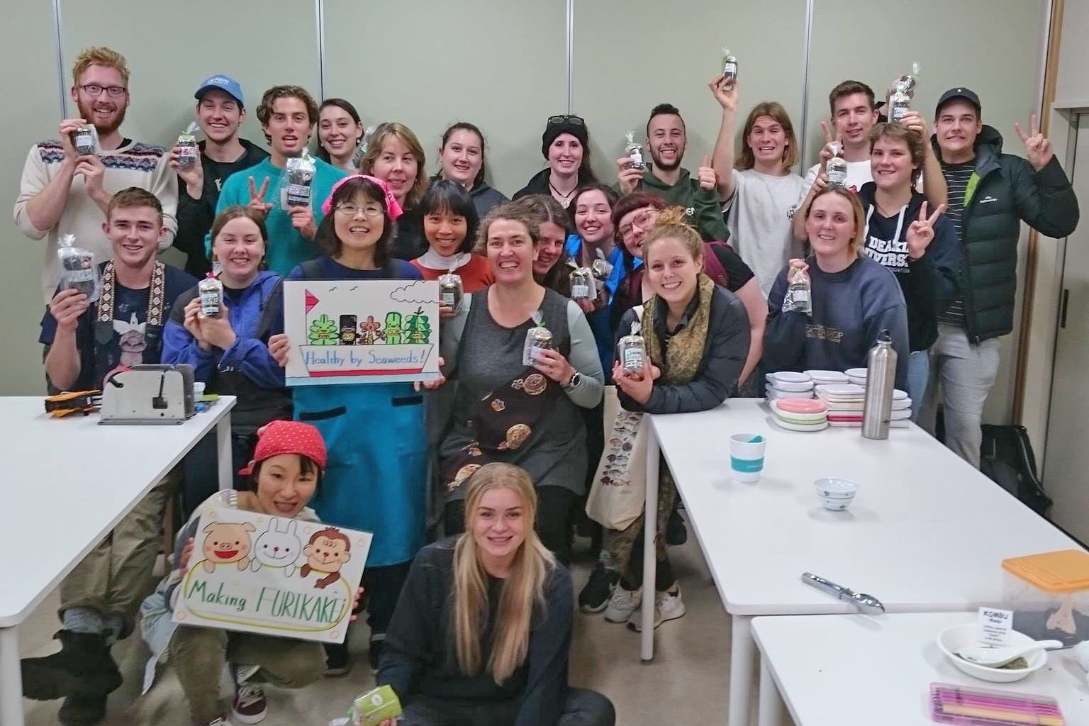 白いテーブルが設置された室内で、2人の日本人の女性が英語とイラストが描かれたボードを持ち、外国人を含む大人数の若者たちが、海藻商品を手に持って笑顔で集合写真を撮っている写真