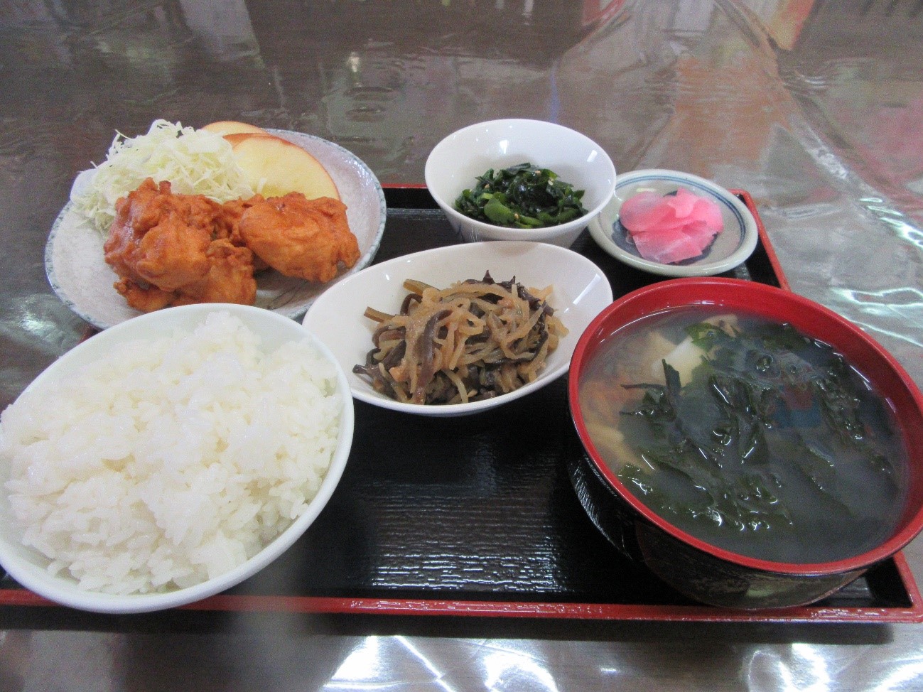 お盆の上に白いご飯、赤い器に入った味噌汁、数個の唐揚げとその横に盛られたキャベツの千切り、小鉢に入ったほうれん草のおひたし、そして彩りのある漬物が整然と並べられている写真