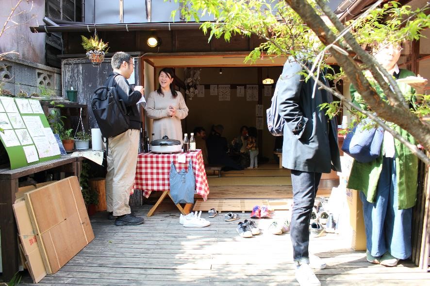 古民家の中では数人が座ってくつろいでおり、木製の床の上で笑顔で立っているベージュの服を着た女性が来場者と話している写真