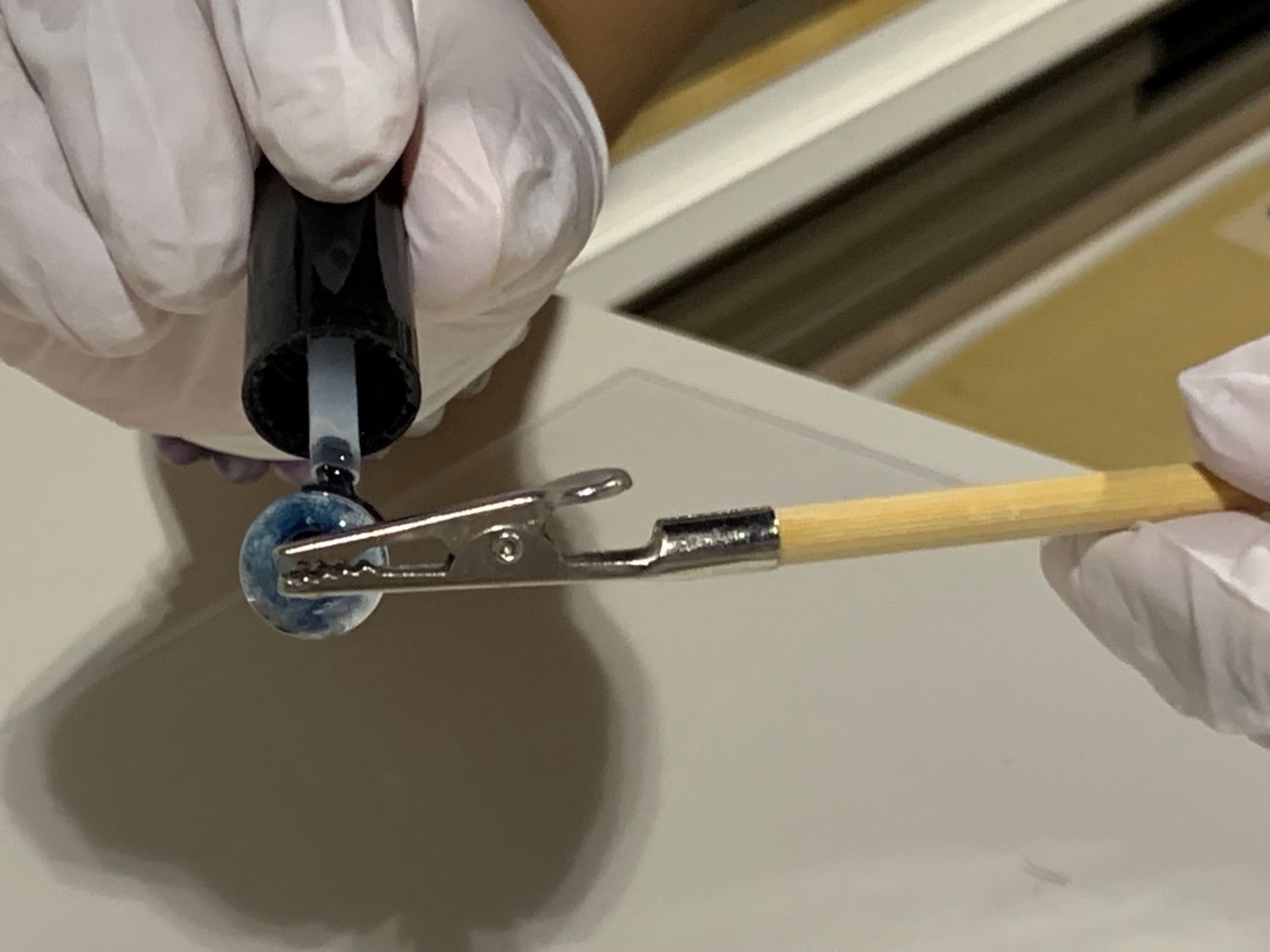 白い手袋を着けた手がピンセット付きの棒で青い球体を固定し、そこにUVレジンを塗っている写真