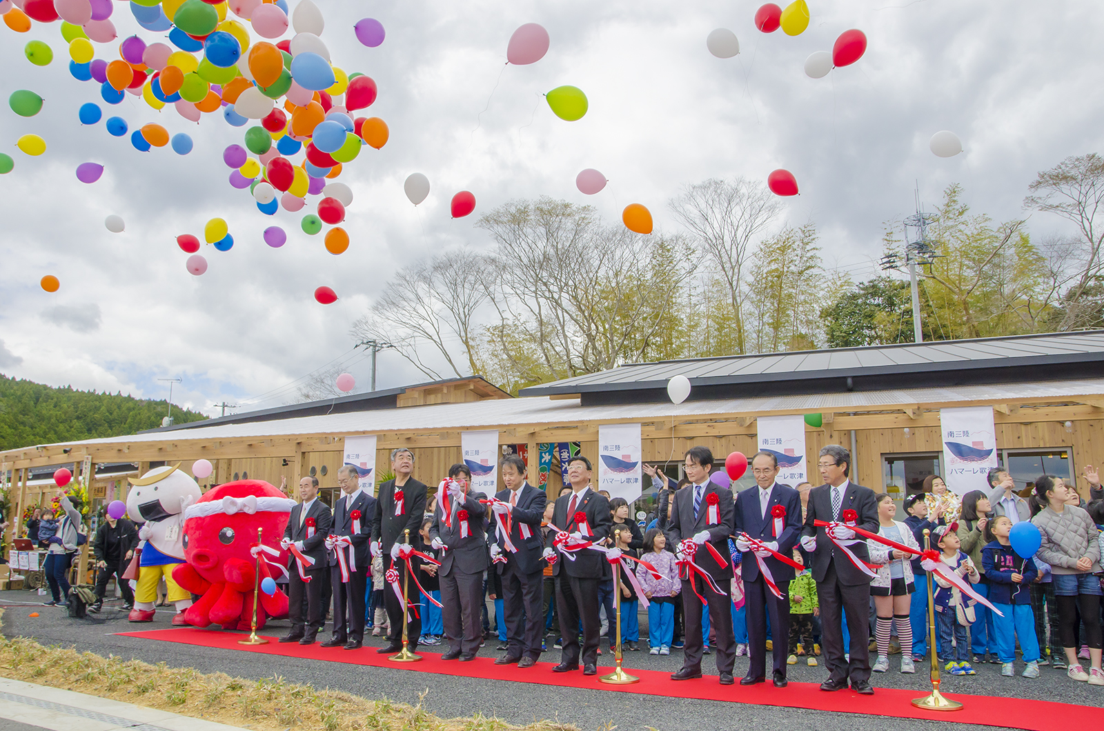 オクトパス君などのキャラクターと関係者が横一列に並んでテープカットが行われ、色とりどりの風船が空に放たれている写真