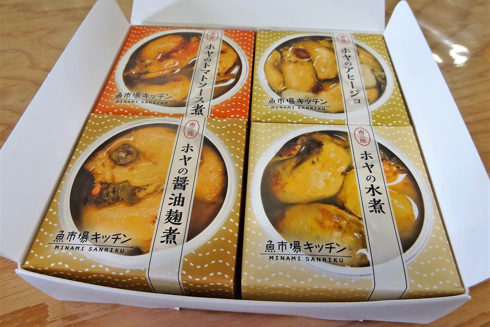 ホヤの缶詰4種類（トマトソース煮、アヒージョ、醤油麹煮、水煮）が箱にきれいに並べられている商品の写真