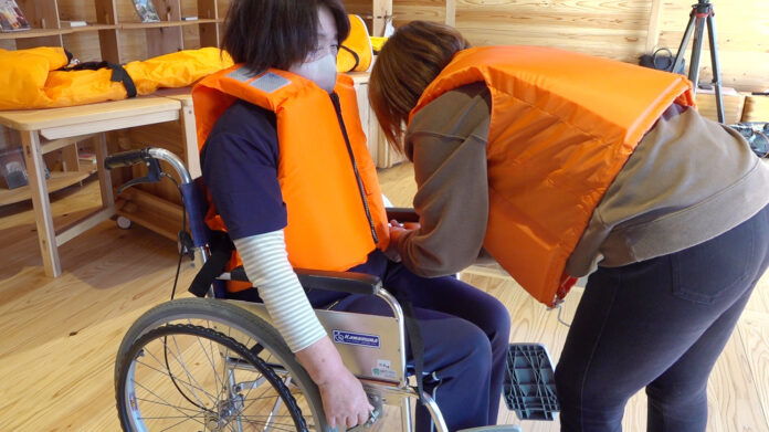 車椅子に座った女性がオレンジ色のライフジャケットを着ており、もう一人がそれを調整している様子の写真