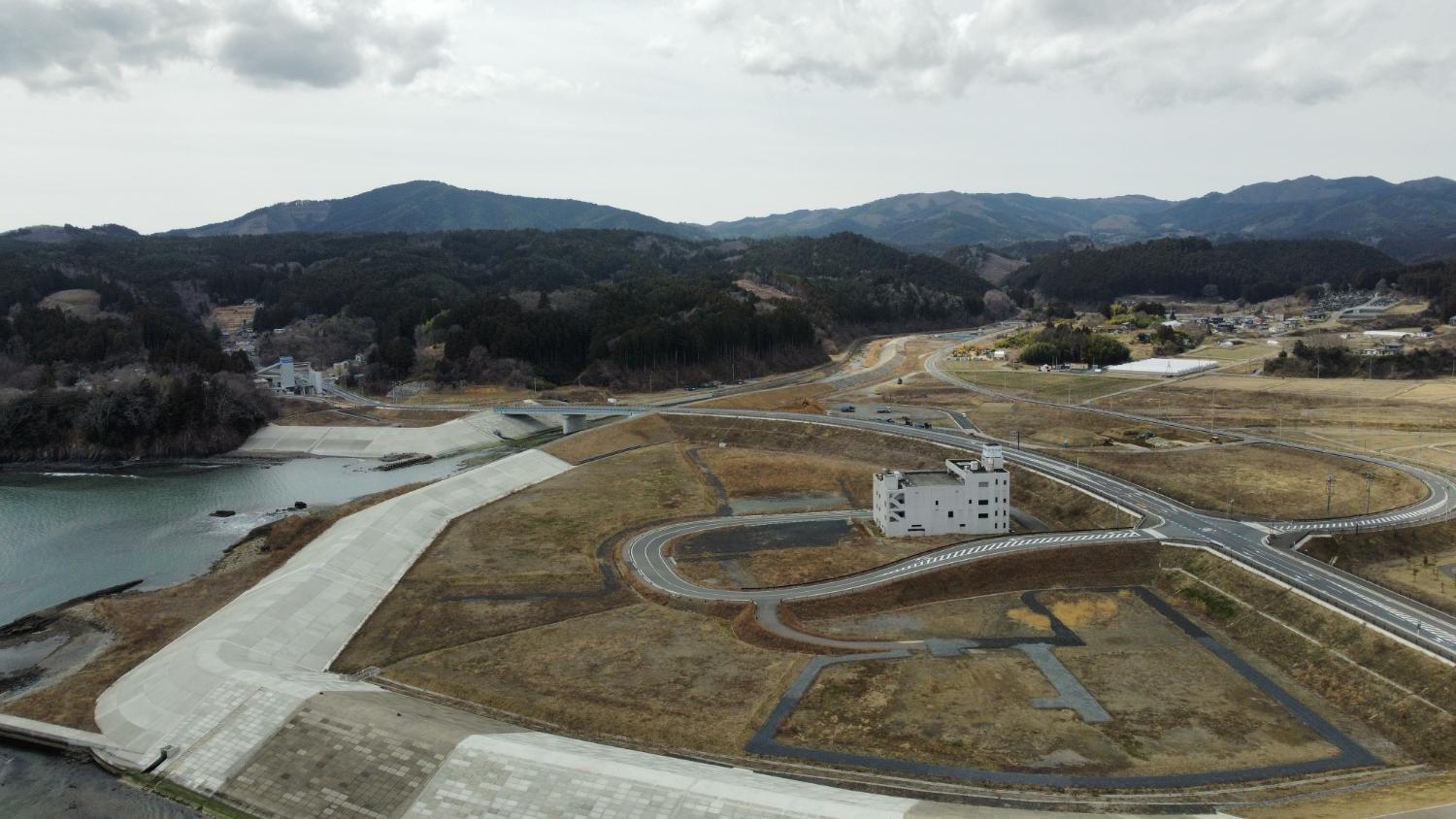 中央には白い建物が立っている広大な造成地の中央を真っすぐに伸びた道路が整備され、背後に山が連なる志津川地区の再開発地の昼間の風景を高台から定点カメラで撮影した写真