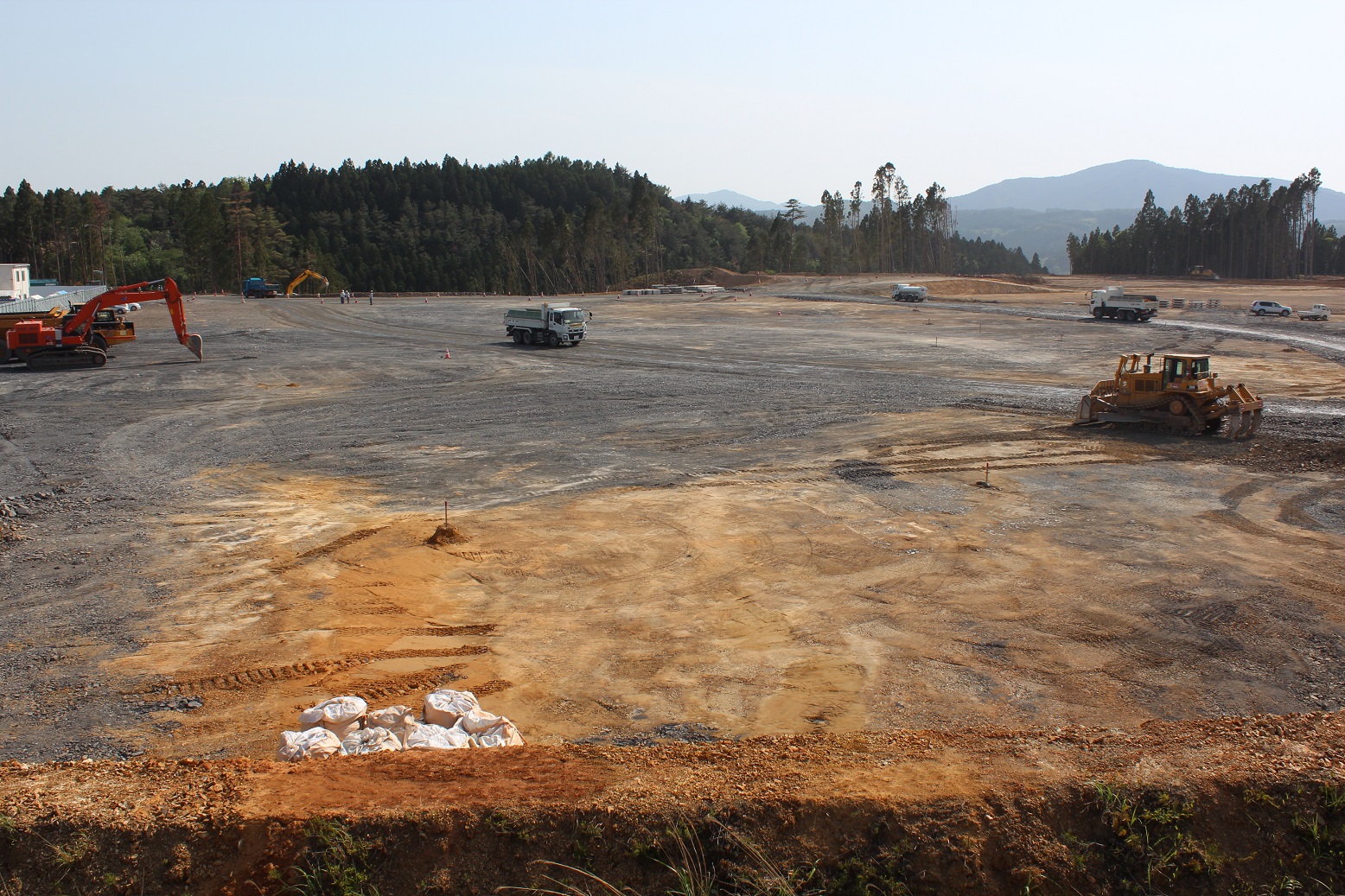 広大な造成地で、ショベルカーやブルドーザー、ダンプトラックが整地作業を行い、地面には砕石が敷かれ、手前に白い土嚢が積まれ、奥には森林と山々が広がっている様子を捉えた写真