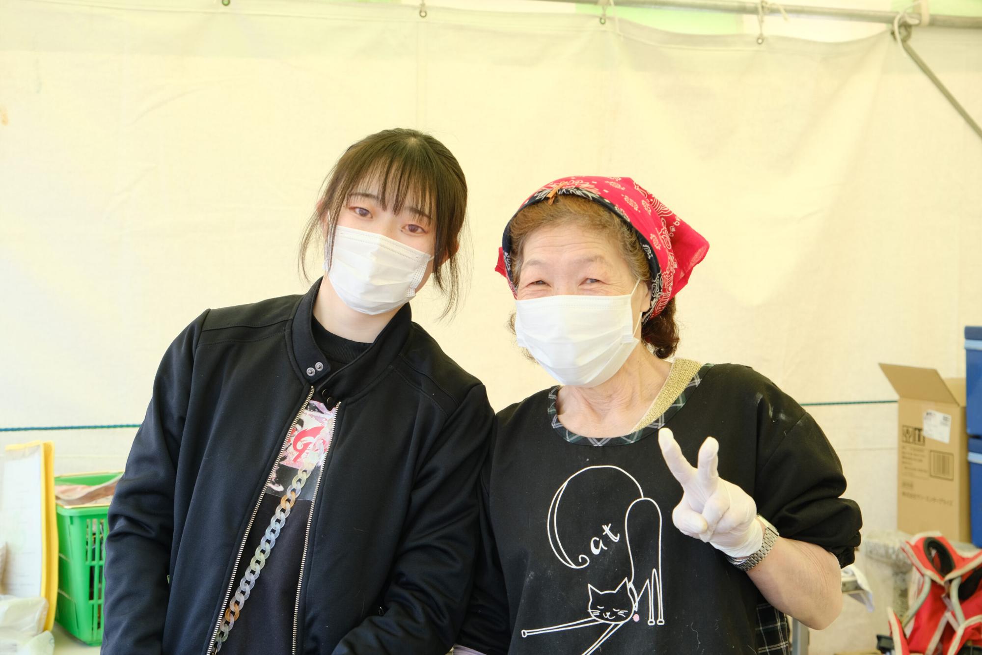 白いマスクをつけた女性と孫の女性が、テントの下でカメラに向かってポーズを取っている写真