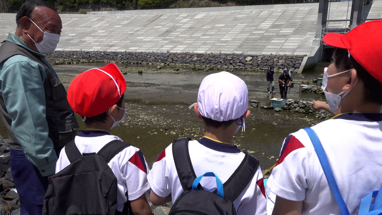 数人の児童が川辺に立ち、年配の男性と話をしながら、遠くで作業している人たちを見ている写真