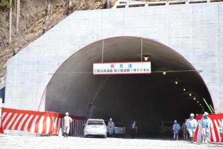 「祝 貫通 三陸沿岸道路 南三陸道路1号トンネル」と書かれた横断幕、両側に紅白幕が掲げられたトンネルの入り口に、ヘルメットを被った複数の人や車両が集まっている写真