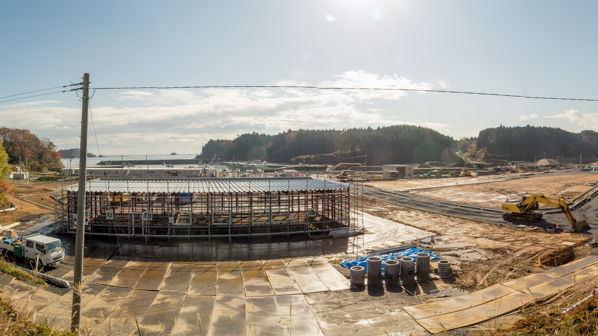 金属の骨組みで部分的に組み立てられた構造物の周囲にはさまざまな建設資材や機材が配置され、左側にはトラックが停車しており、右側ではショベルカーが作業を行っている建設現場の写真