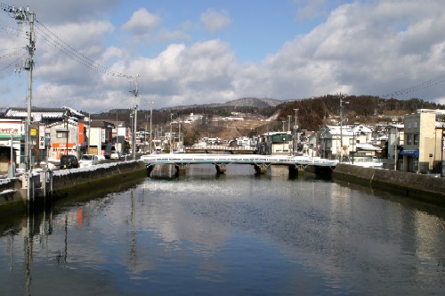 水面が静かに空を映す運河のような川の正面に、橋が架かり、両岸には雪化粧をした民家や商店、そして奥には雪の積もった山が見える冬の街並みの写真