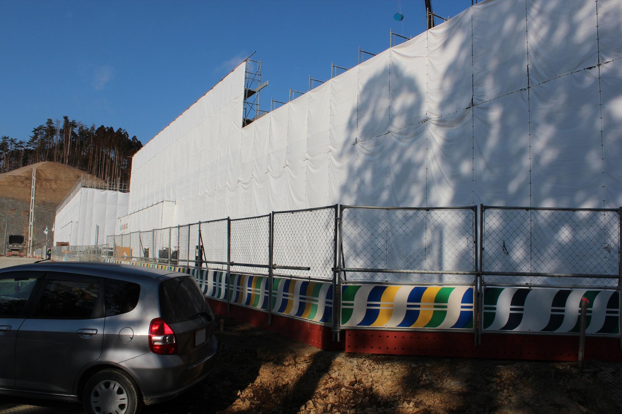白いシートで覆われた建設現場の外壁が日差しを受けて明るく映え、手前には車両とカラフルな模様入りの仮囲いフェンスが並んでいる写真