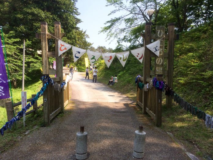 山間の小道の入り口にある木製の門に、ひらがなで「ひころマルシェ」と書かれたフラッグガーランドが掲げられ、人々がのんびりと歩いている様子の写真