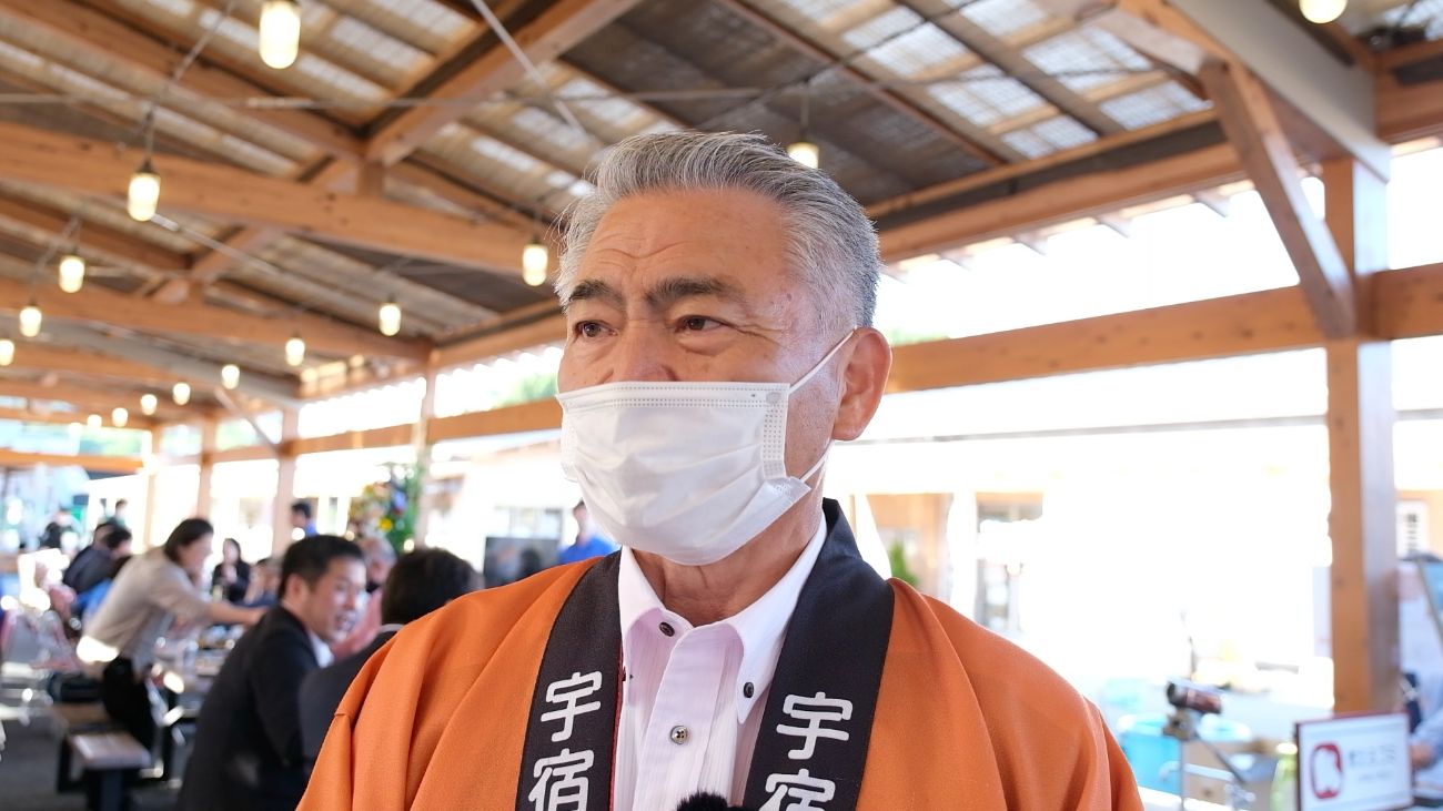 復興市の屋台の下で橙色の法被を着た河井達志さんが、白いマスクをつけてインタビューに答えている写真