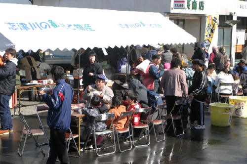 路上に白いテントが張られ、その下で多くの人々がテーブルとパイプ椅子に座って食事を楽しんでいる、寒鱈祭りの様子の写真