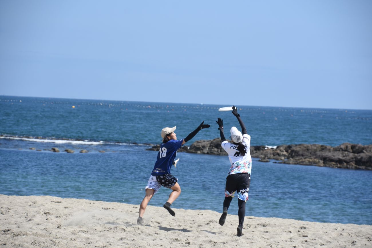 海辺で2人の選手がディスクを奪い合うように両手を伸ばして跳び上がっている様子の写真