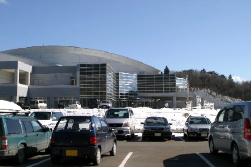 雪の積もった広い駐車場に車が駐車され、その奥にはドーム型の屋根とガラス張りの壁面を持つ近代的な大型公共施設が建っているベイサイドアリーナの風景写真