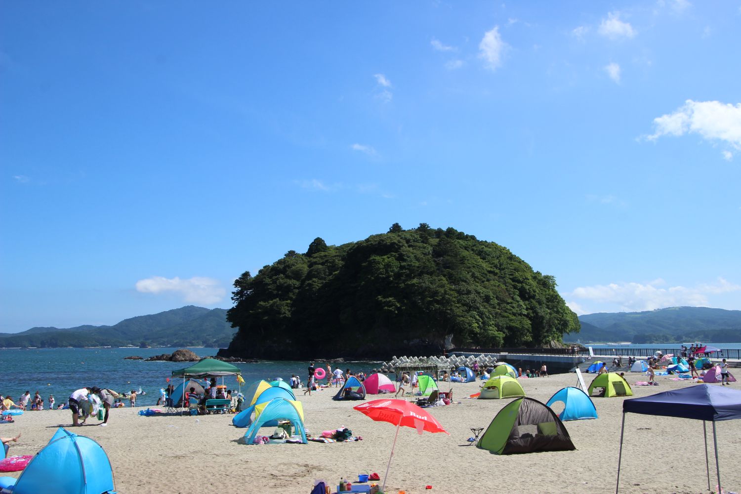 青空の下、砂浜に並ぶカラフルなテントやパラソルの間で多くの人々が海水浴を楽しみ、背景には緑豊かな荒島が見える夏の海辺の写真