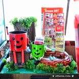 赤いバケツ型キャラクターが右手を上げて木に座り、傍に緑のバケツ型キャラクターが座った、林をイメージしたパンフレットスペースが展示されている写真