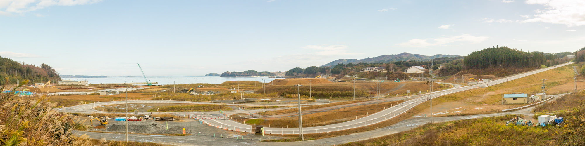 白いガードレールが設置された曲がりくねった道路が複数あり、建設車両や機材が点在し背景には水辺が広がり、遠くには陸地や島が見え、周囲には草地や丘が広がり、遠くには木々が茂っている工事中の海沿いの地域をパノラマで写した写真