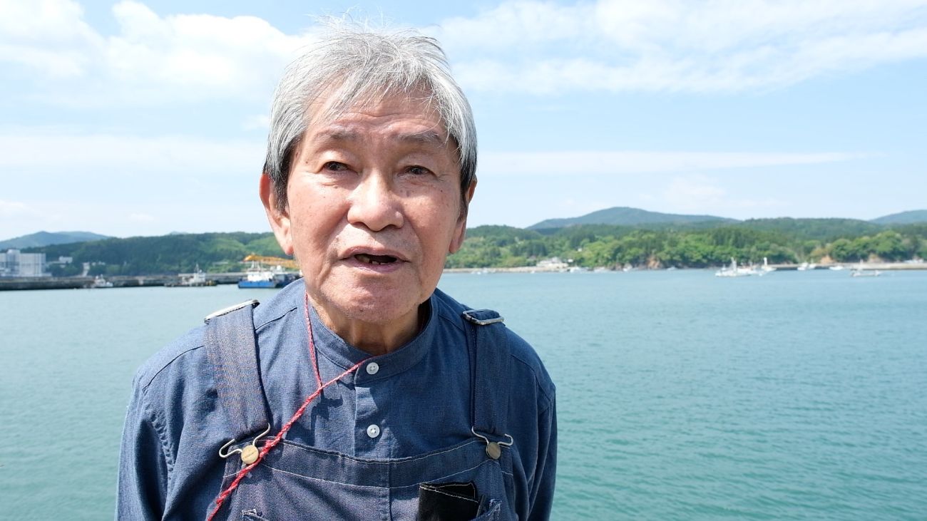 港を背景にした屋外で作業着を着た男性が、復興について語っている写真