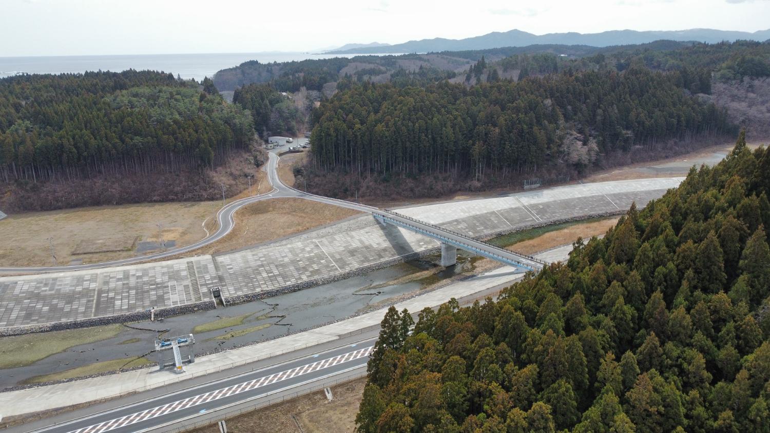 コンクリート造りの高い護岸に挟まれた川の上に橋がかかっており、その奥に山に囲まれたカーブの多い道と住宅が点在している静かな山間の風景で、画面右手前には杉の木が密集している、整備された堤防と橋の昼間の風景を高台から定点カメラで撮影した写真