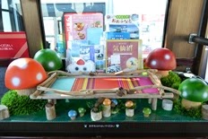 大小様々な大きさのカラフルなキノコのフィギュアが、流木に囲まれた縞模様の布が敷かれた小さな木の橋、芝生風の緑の台の上に作られ囲まれたパンフレットスペースの写真