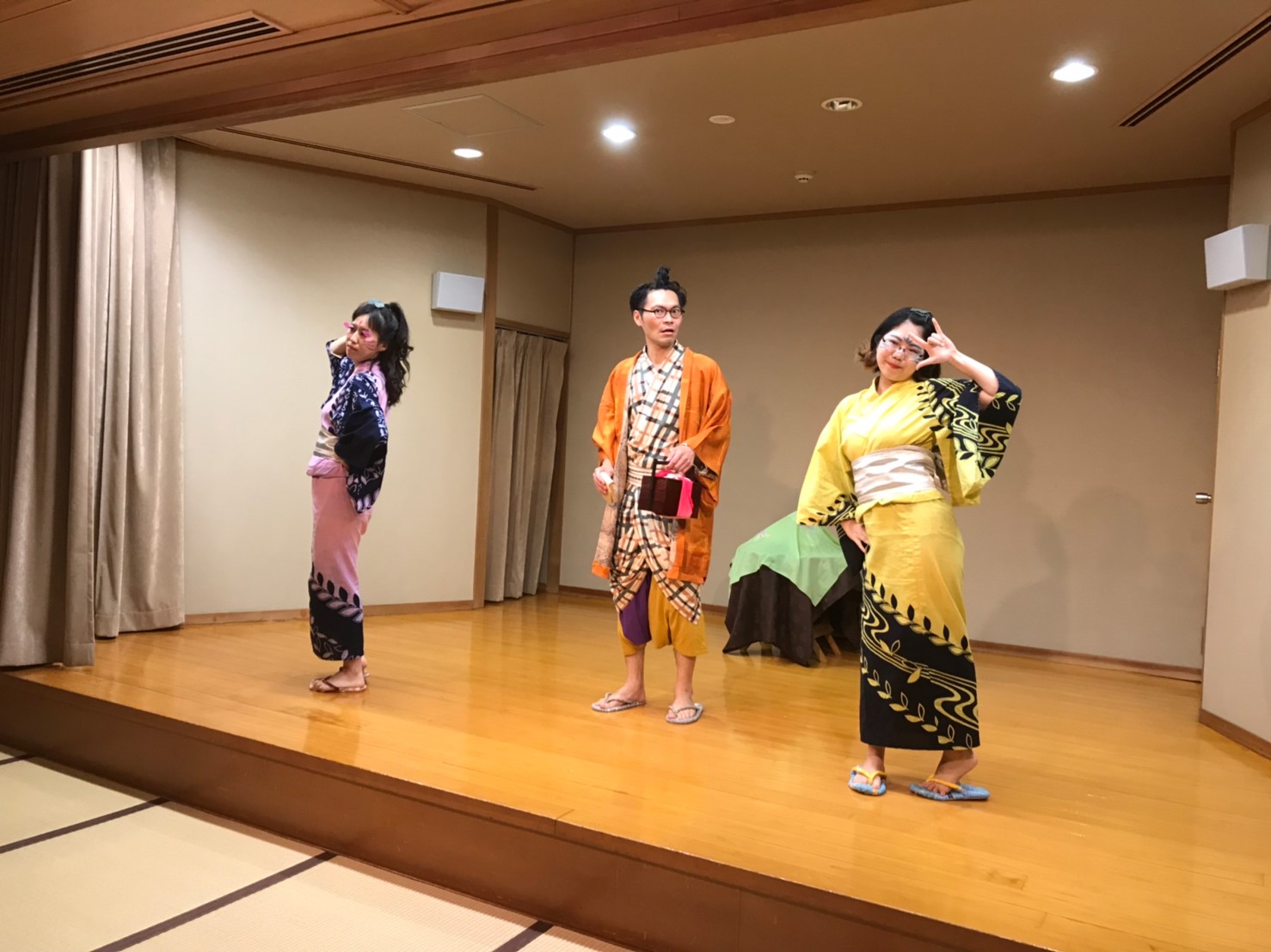 舞台上で黒い柄の入ったピンクと黄色の衣装を着た2人の女性と、中央にオレンジ色の羽織を着た男性が立ち稽古をしている最中の写真