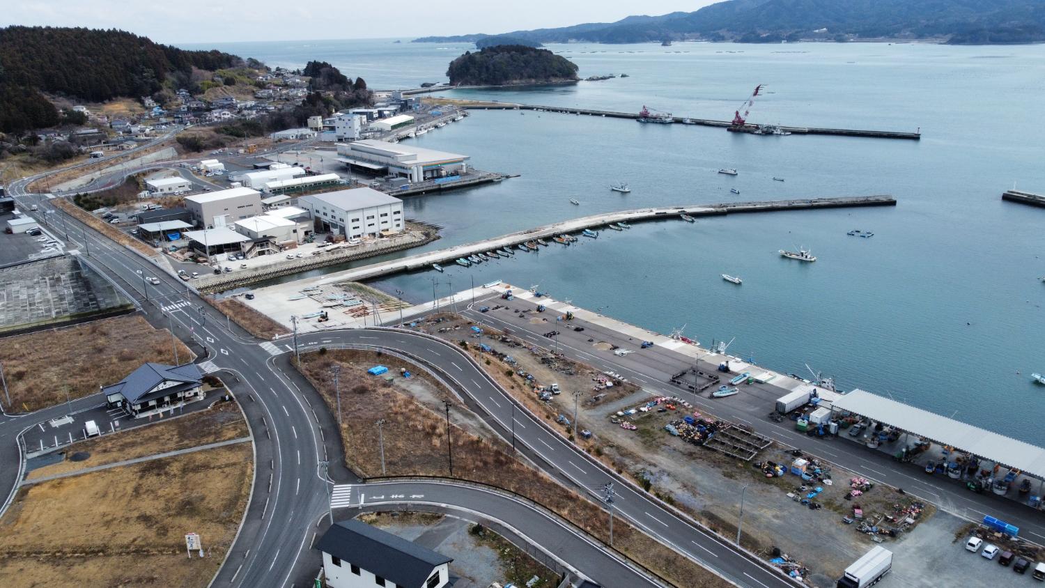 湾内に多くの漁船が浮かぶ漁港を中心に、奥には山に囲まれた街並みが広がり、整備された建物や道路が見える港周辺の昼間の風景を高台から定点カメラで撮影した写真